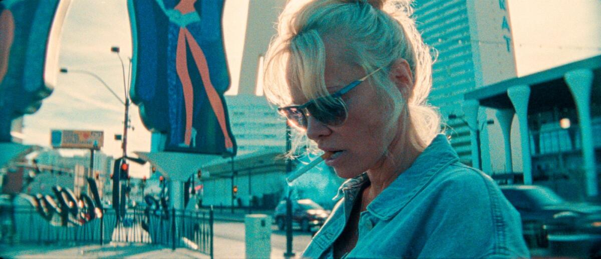 Pamela Anderson stands outside looking down with a cigarette in her mouth.