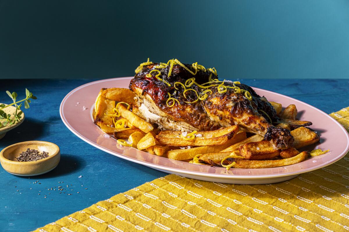Closeup of an oblong pink plate on a table with blue and gold cloths. Cooked chicken sits atop French fries. 