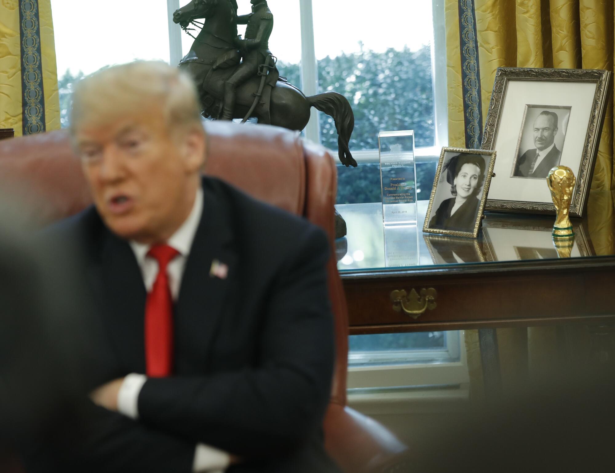 Um homem de terno escuro e gravata vermelha, de braços cruzados, fala em frente a uma mesa com fotos emolduradas e uma estátua de uma pessoa a cavalo