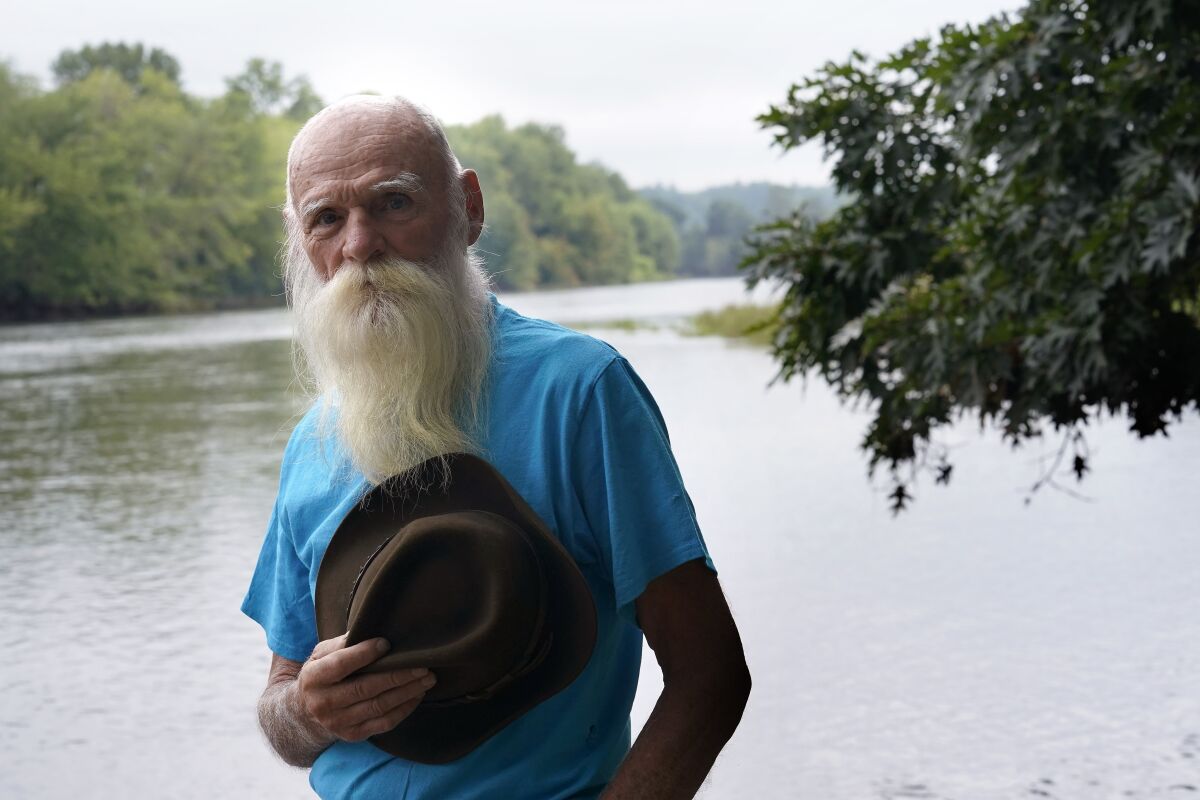 FILE — David Lidstone, 81, stands for a photograph near the Merrimack River, Tuesday, Aug. 10, 2021, in Boscawen, N.H. Lidstone, a former hermit in New Hampshire, known to locals as "River Dave," whose cabin in the woods burned down after nearly three decades on the property that he was ordered to leave, and who received more than $200,000 in donations, has been charged with trespassing there once again. Lidstone still disputes that he is on the property, and was arrested on a trespassing charge on Dec. 14, 2021. He faces a court hearing in March of 2022. (AP Photo/Steven Senne, File)