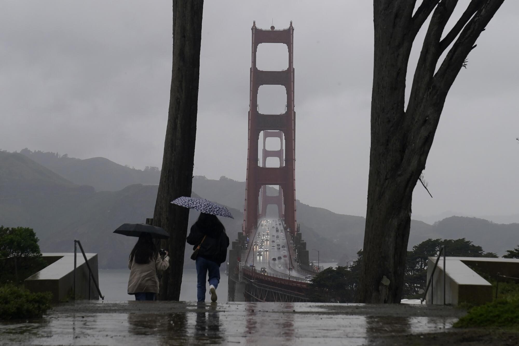 Two people hold umbrellas as the walk in the rain. The Golden Gate Bridge rises in the background.
