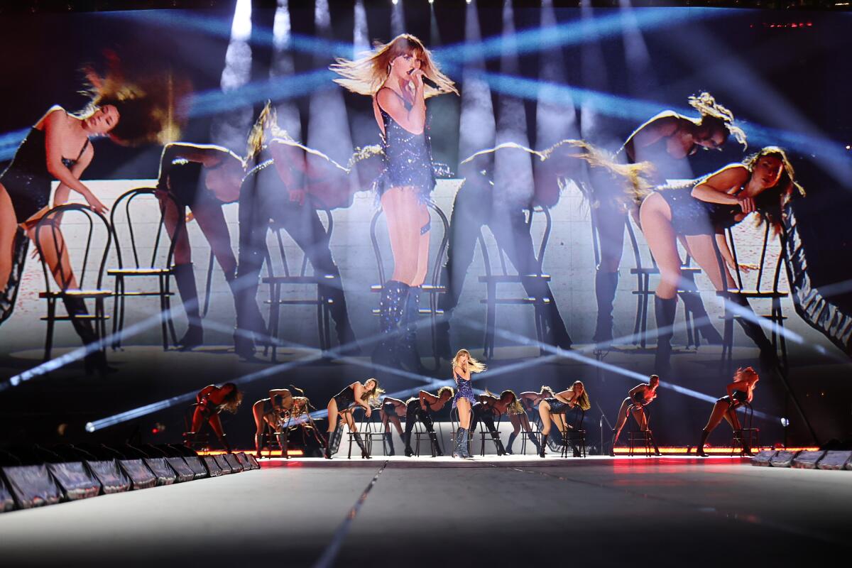 Taylor Swift in a sparkling bodysuit and boots sings in a microphone. Backup dancers use chairs as props.