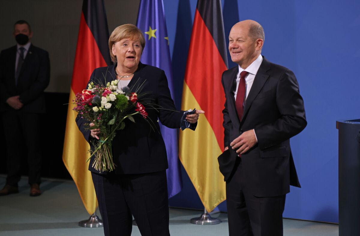 A woman in a dark suit, left, holding a red-and-white bouquet of flowers gestures to a smiling man standing beside her