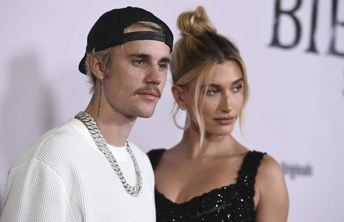 Justin Bieber in a white thermal, black ballcap and silver chain posing with Hailey Baldwin in a black top and updo