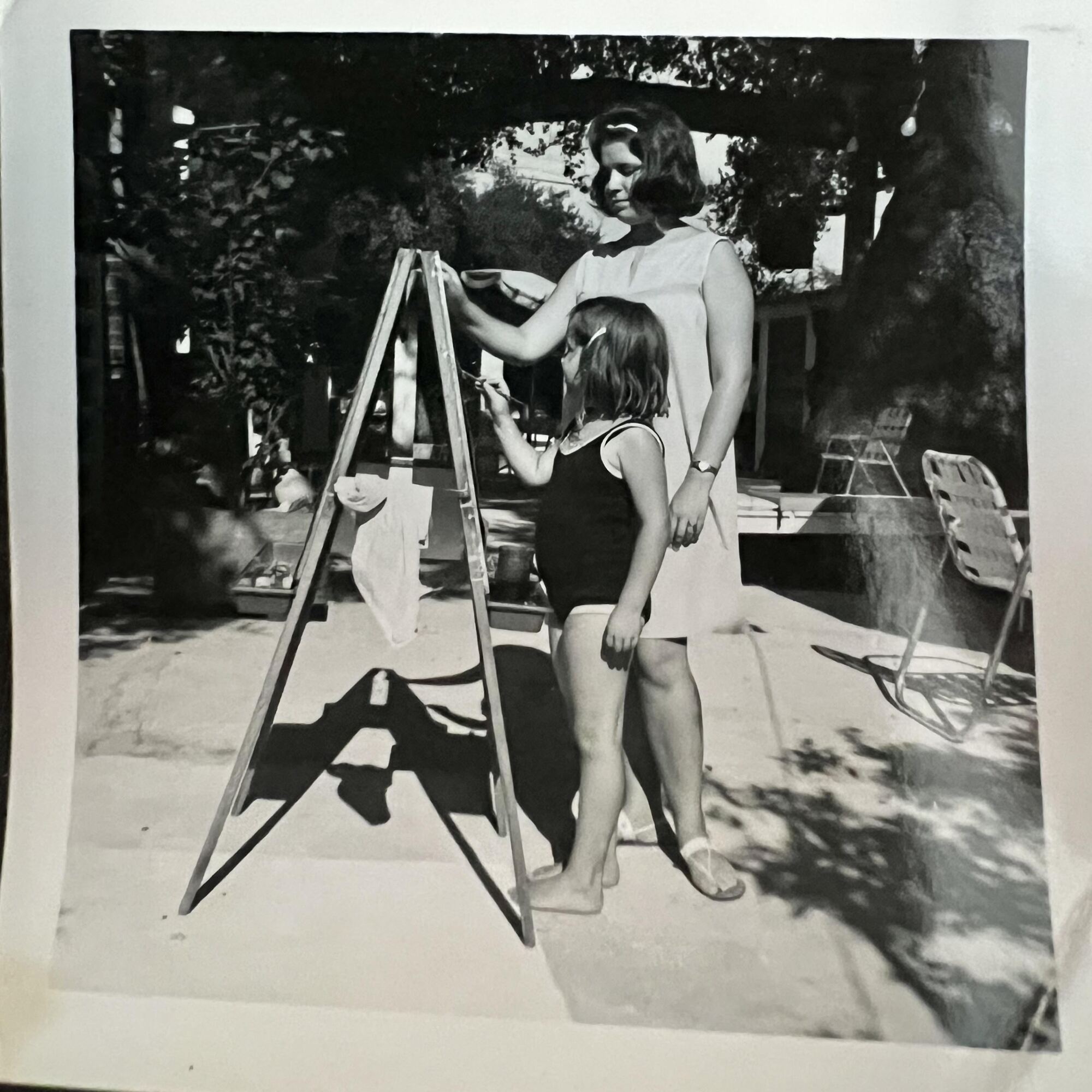 A young girl and a teenage girl stand together, painting, at an easel 