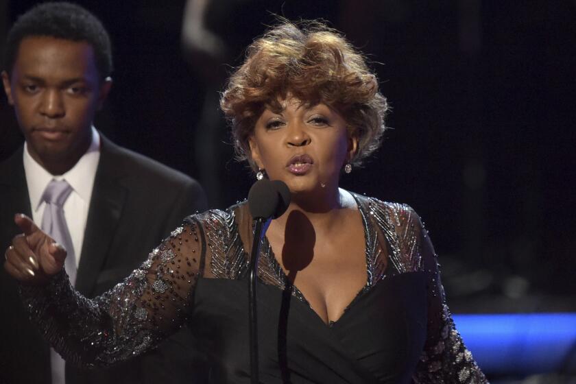 A woman with short hair wearing a black gown speaks and points onstage behind a microphone