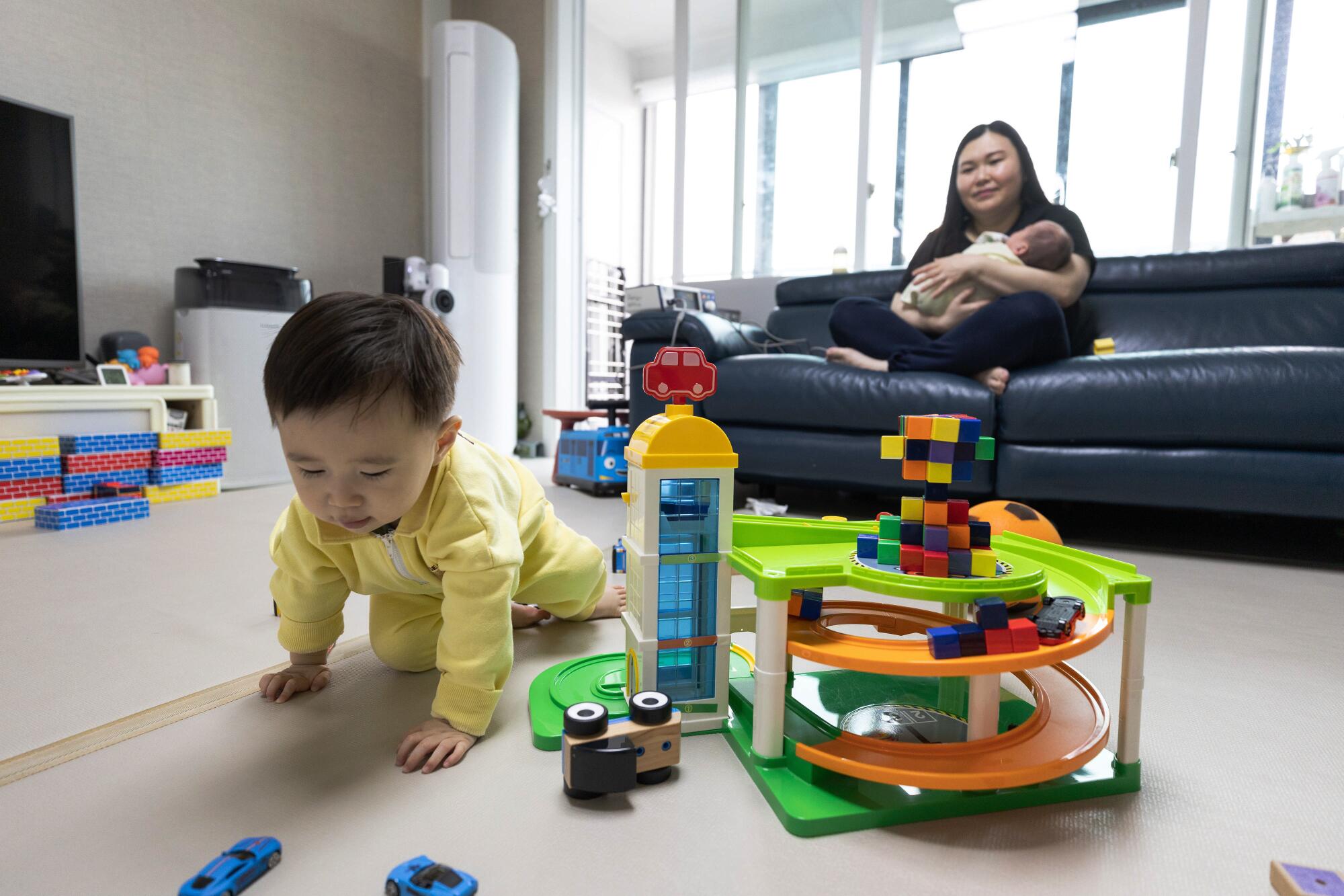 A woman sits on a couch holding an infant while a baby crawls amid toys in the foreground. 