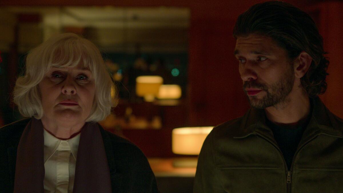 A white-haired woman standing next to a dark-clad man who looks toward her.
