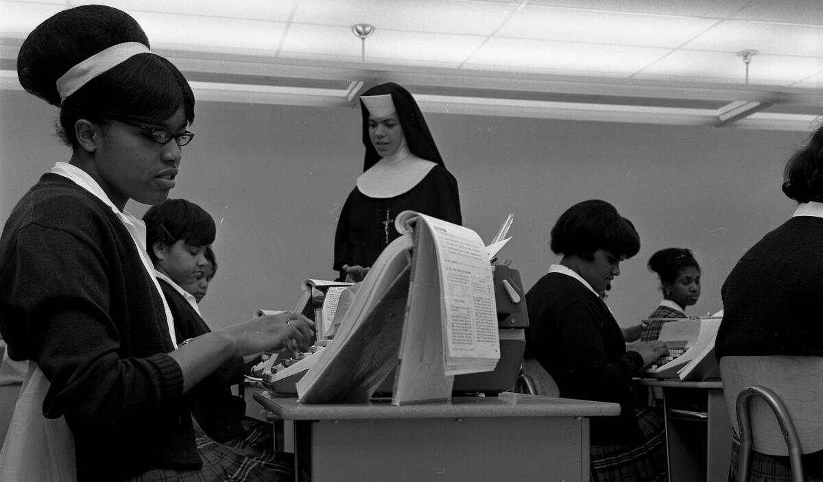 Girls sit at typewriters in a classroom, with a nun in the background.