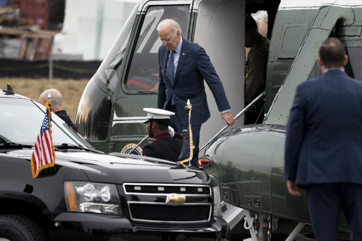 A man with gray hair, in dark suit and blue tie, steps from a helicopter.