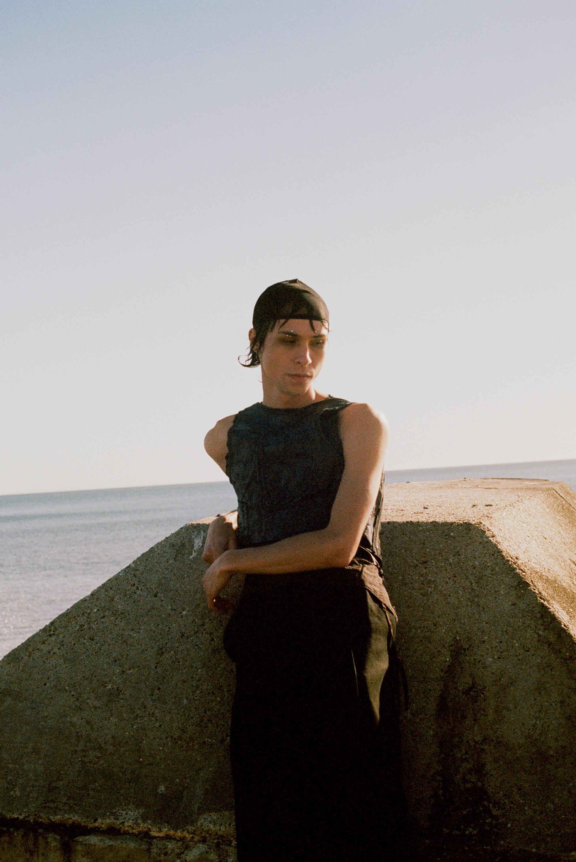 A model in a black tank top leans against a concrete slab on the beach.