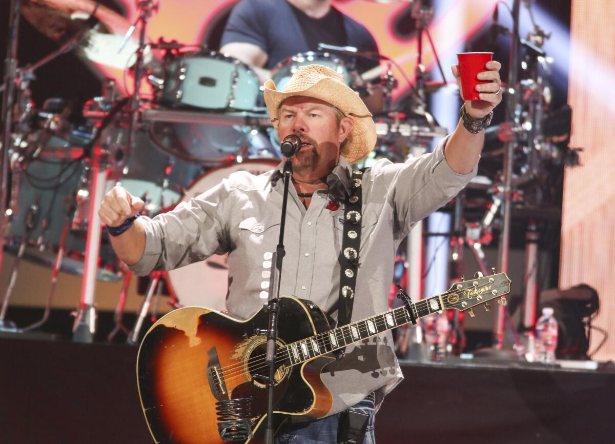 A man in a cowboy hat holds a red solo cup and plays guitar while singing into a microphone