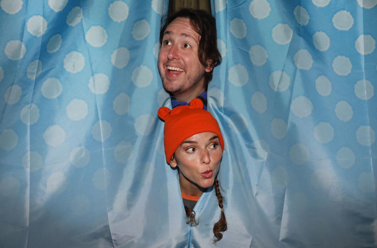 A smiling man and a woman in a beanie with cat ears pop their heads out of a polka dot curtain. 