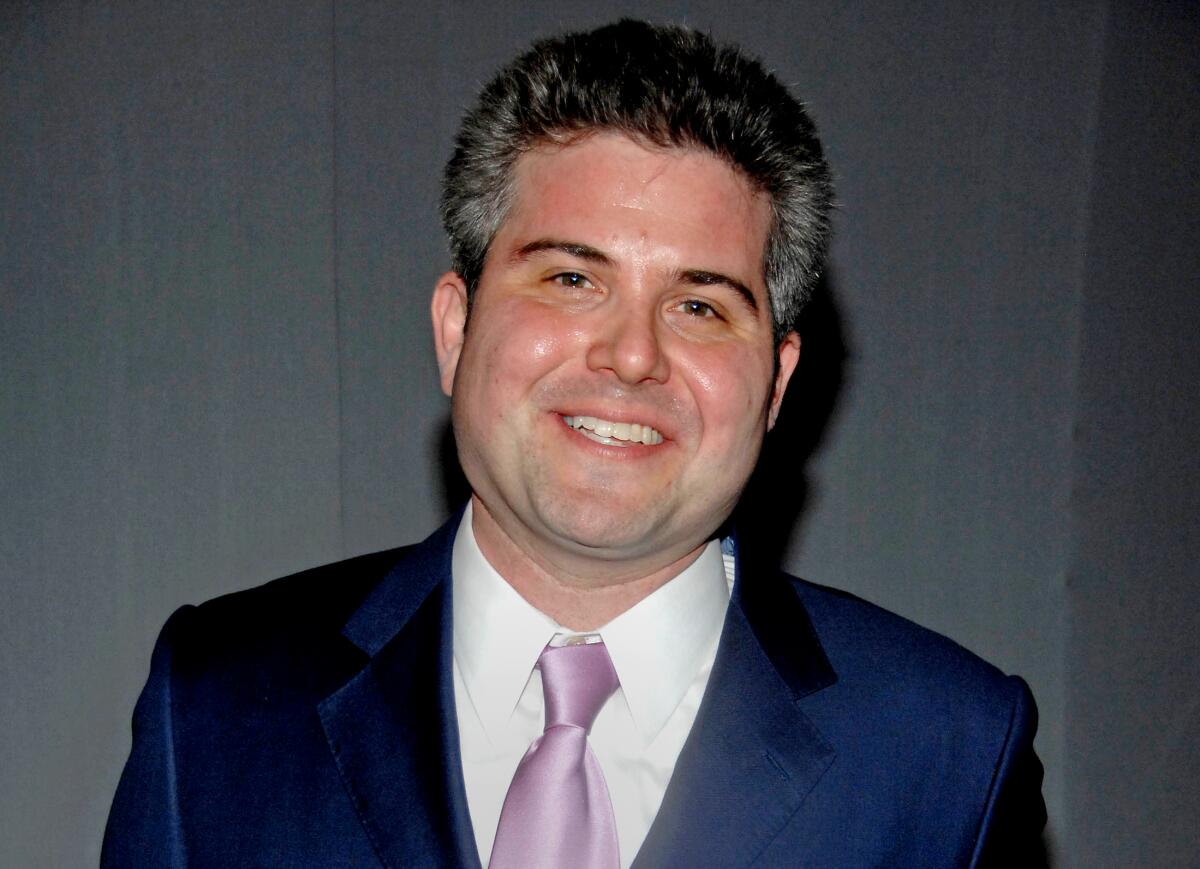 Adam Epstein in a blue suit with a pink tie smiling against a gray backdrop.