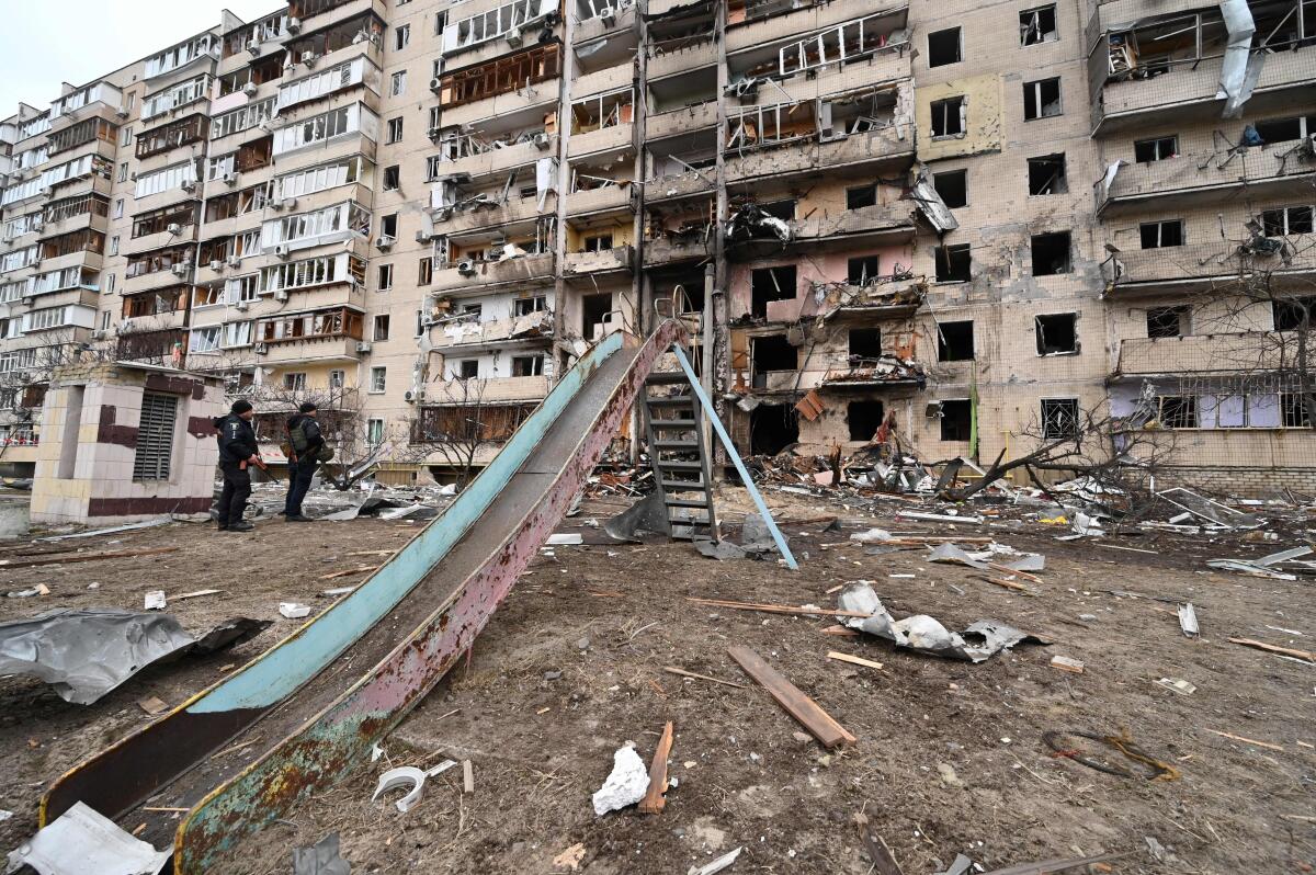 Police officers guard a large damaged building in Ukraine. A playground slide is in the foreground.

