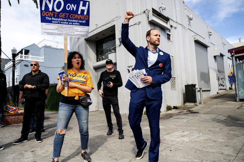 A man in a suit raises his right fist as he walks past a woman holding a sign that reads "No on H"
