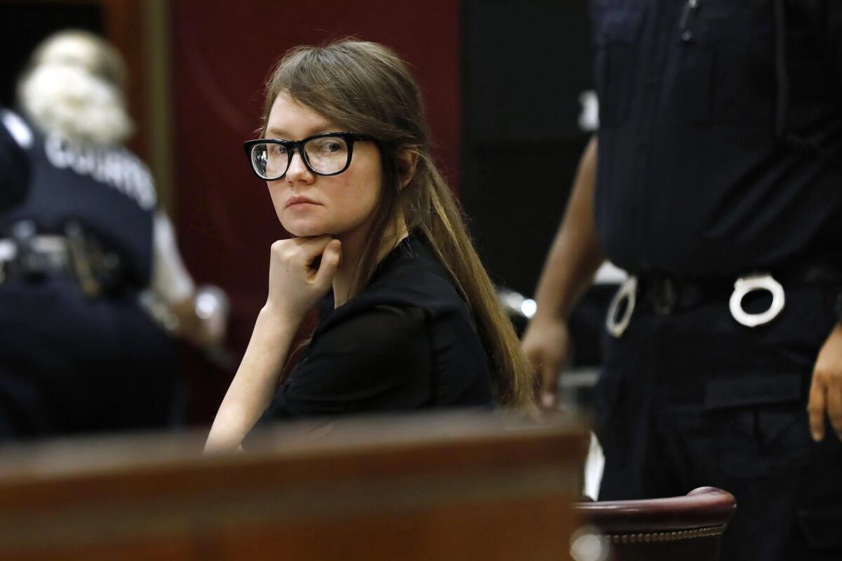 A woman in glasses sits at a courtroom defense table with her chin on her hand