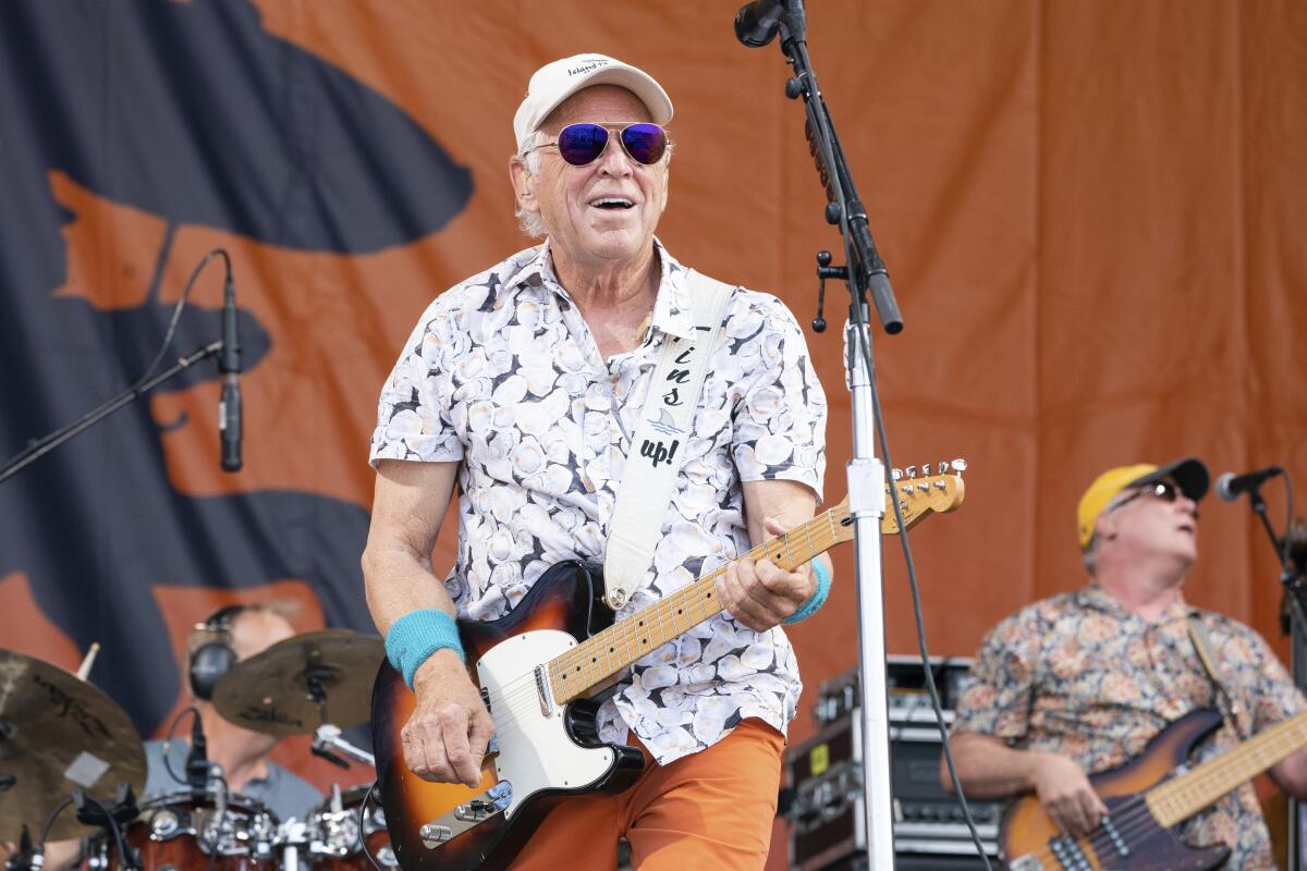Jimmy Buffett performs while wearing flowered shirt, cap and sunglasses and strumming electric guitar on stage. 