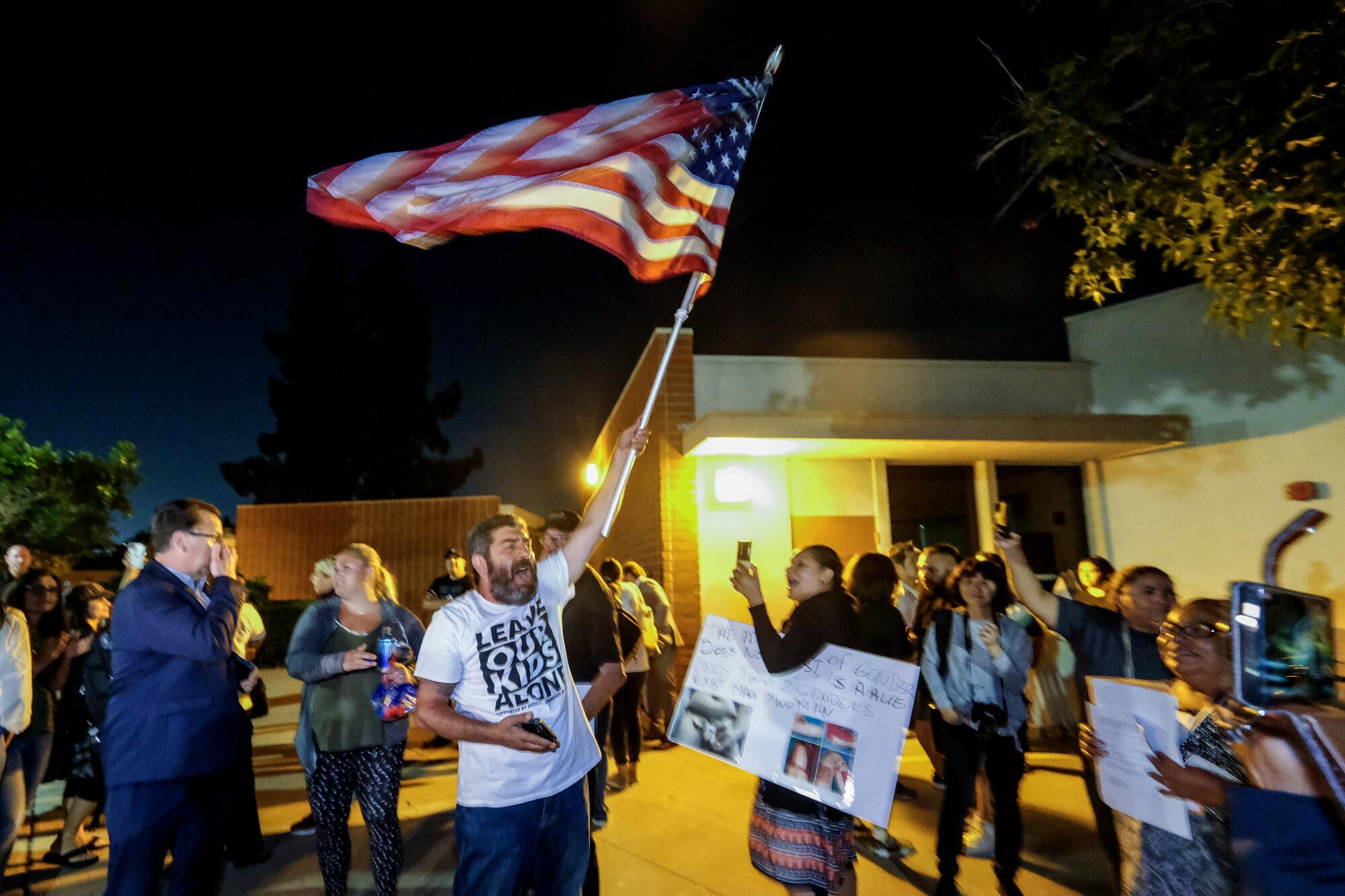 A man waves an American flag outside a building as others stand, some with phones raised.
