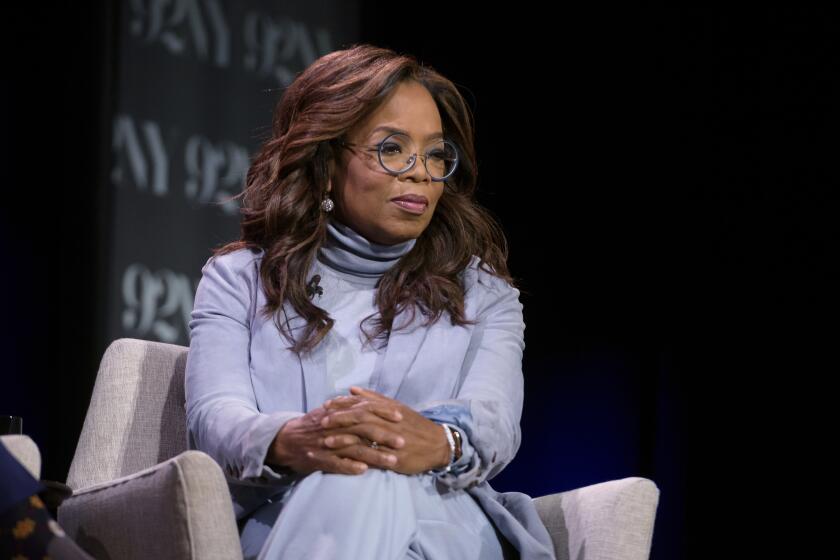 Oprah Winfrey sits on a chair with her legs crossed and her hands folded over her knees.