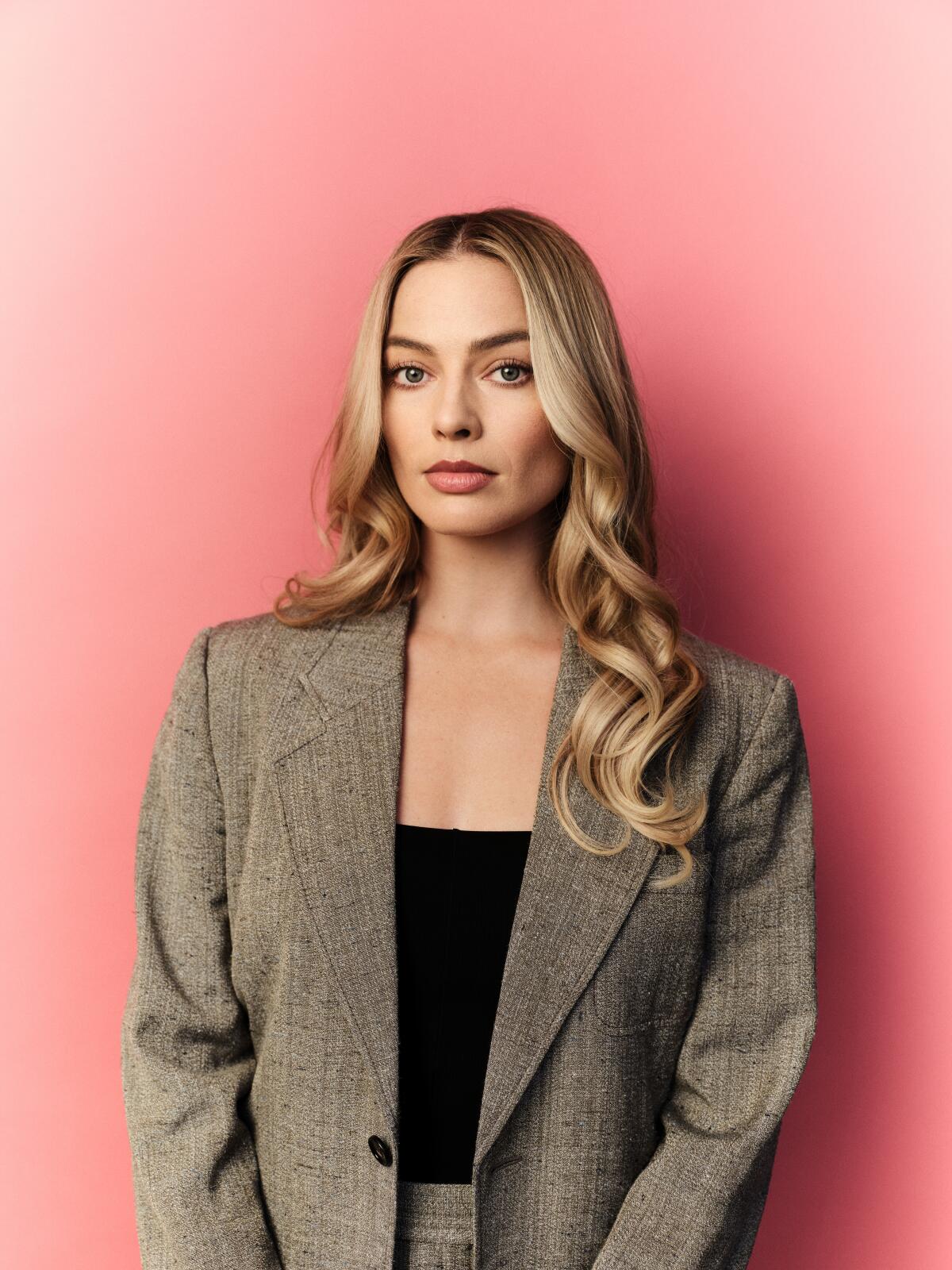 Margot Robbie, with her long blond hair in waves, stands in front of a pink background.