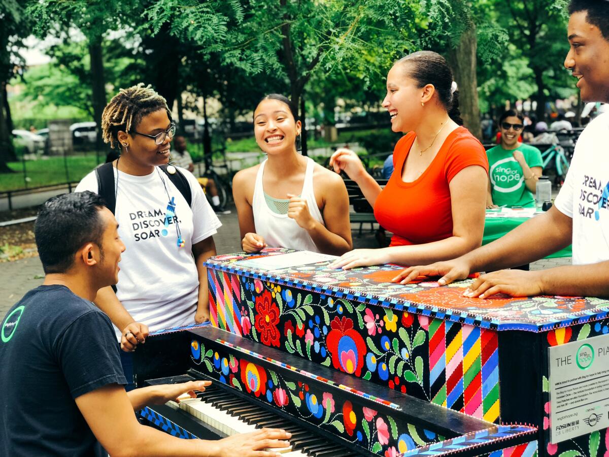 Young people gather around a brightly painted piano in a public park