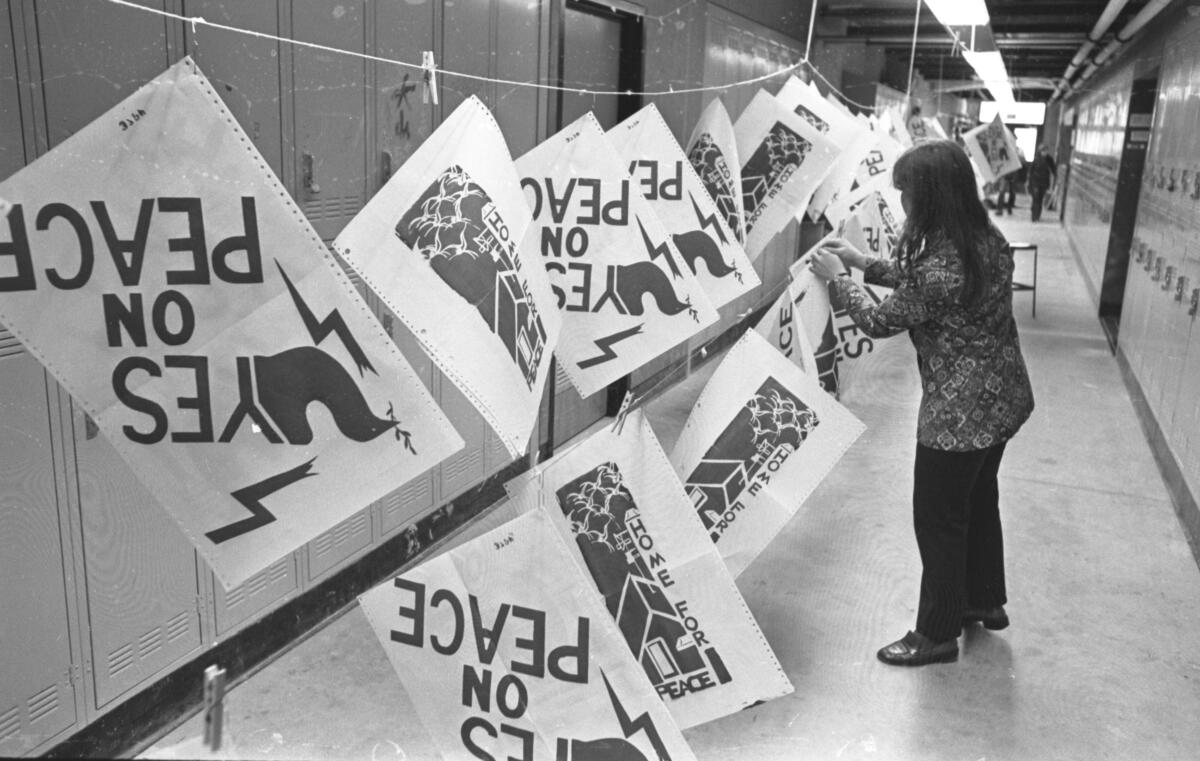 A person stands in a hallway lined with lockers. Clotheslines hold posters that read "Yes on Peace."