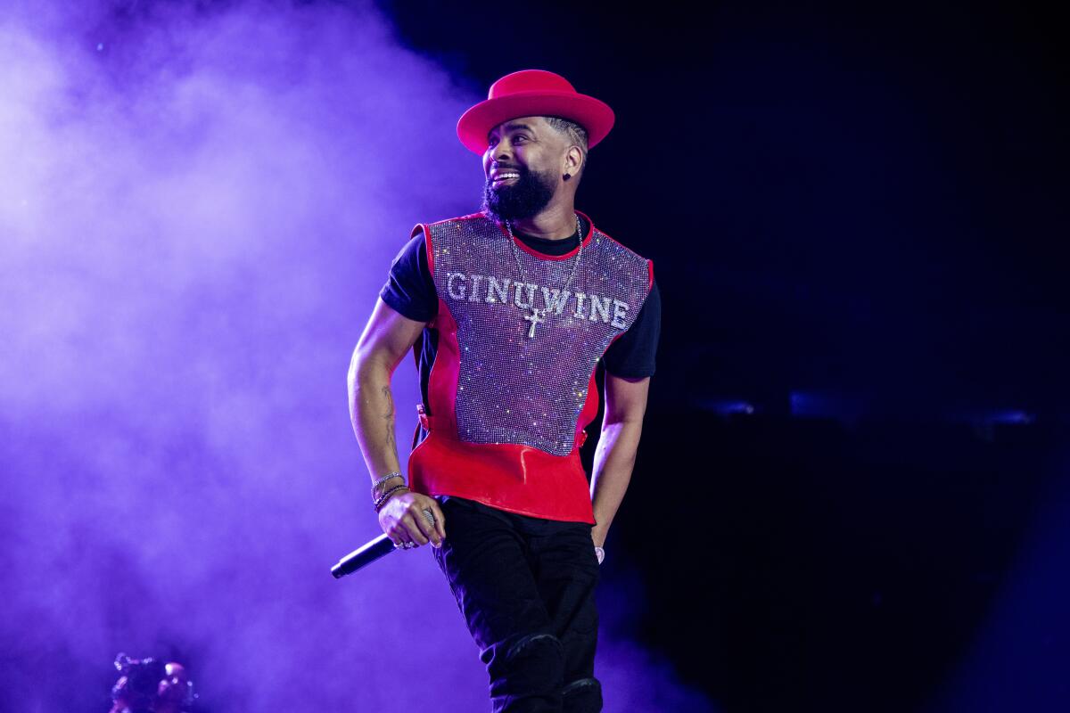 A man wearing a shirt that says Ginuwine and a red hat to match smile whiles holding a microphone on stage