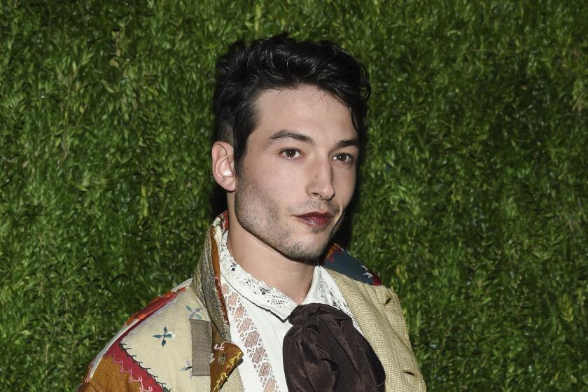 A person with short dark hair, stubbly facial hair and dark lipstick poses in front of foliage