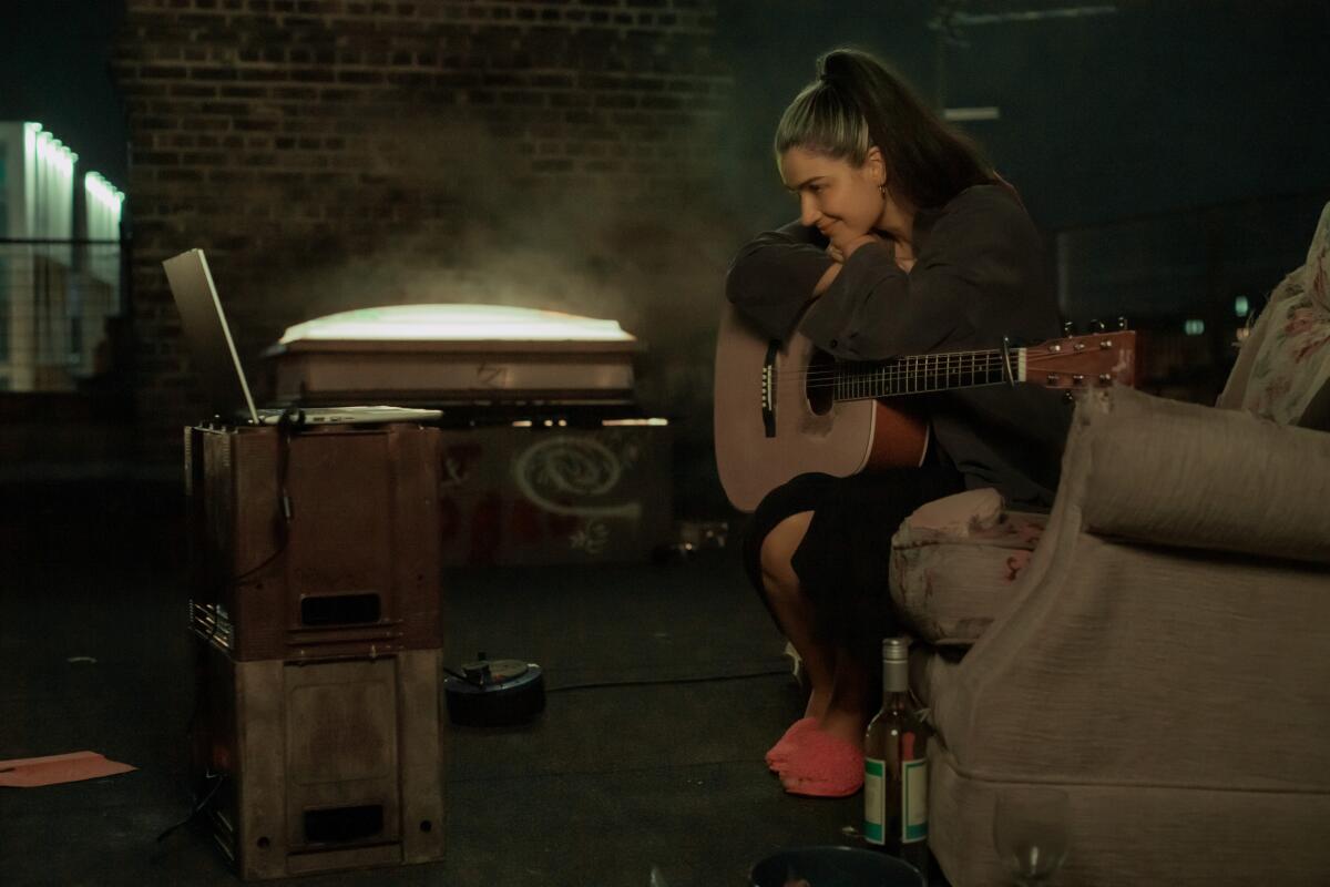 A woman with a guitar sits on a couch looking at a laptop.