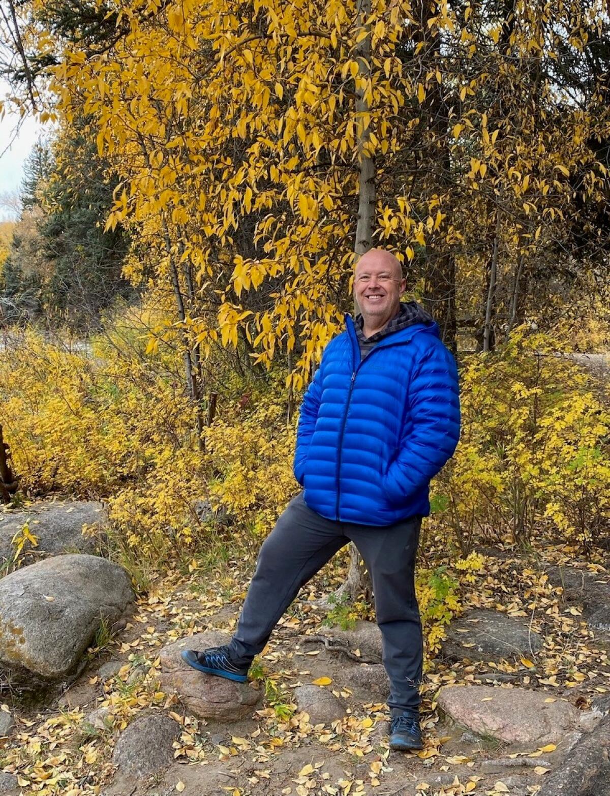 A man in a blue jacket stands in front of some trees