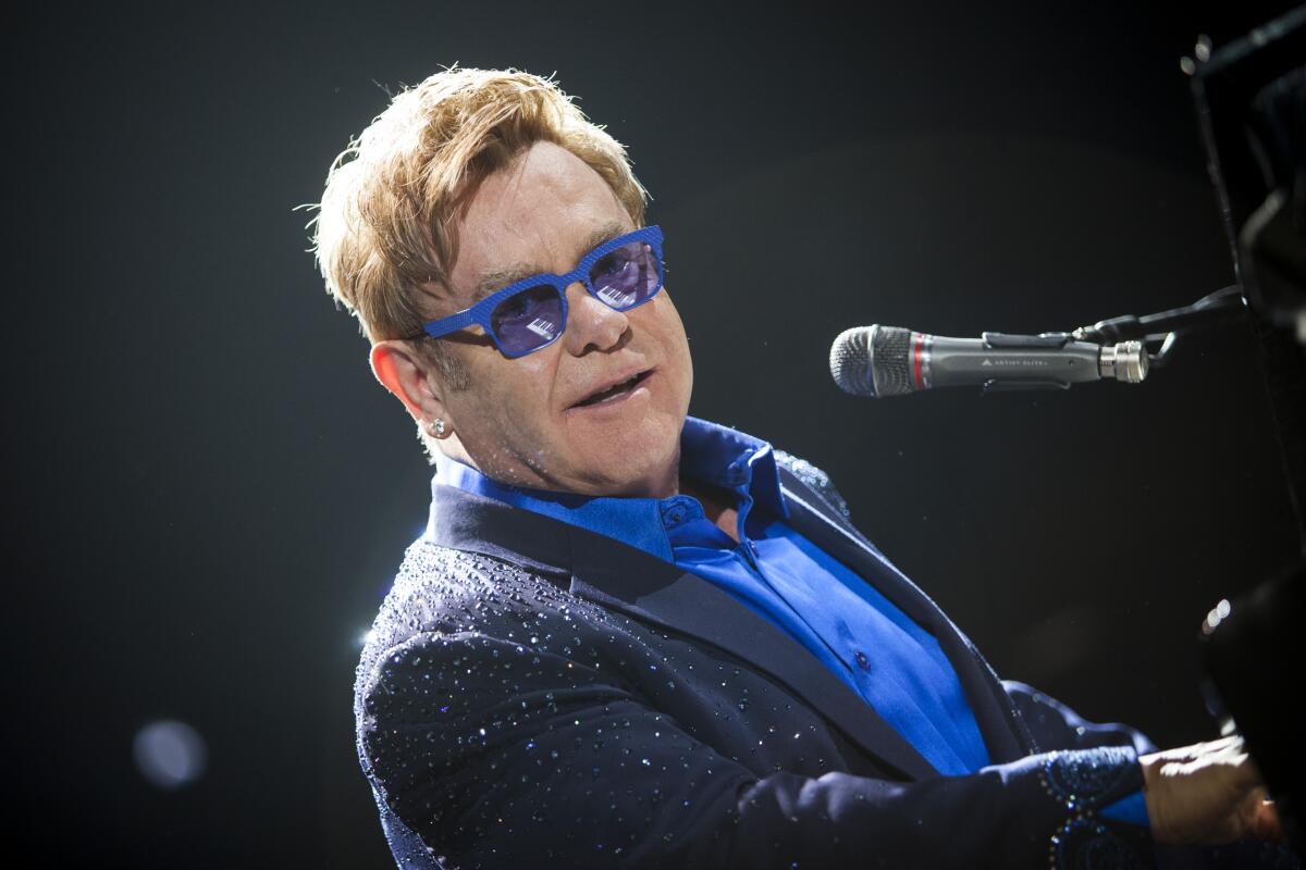 Elton John wears blue glasses to match a blue shirt as he leans back while playing the piano and singing onstage