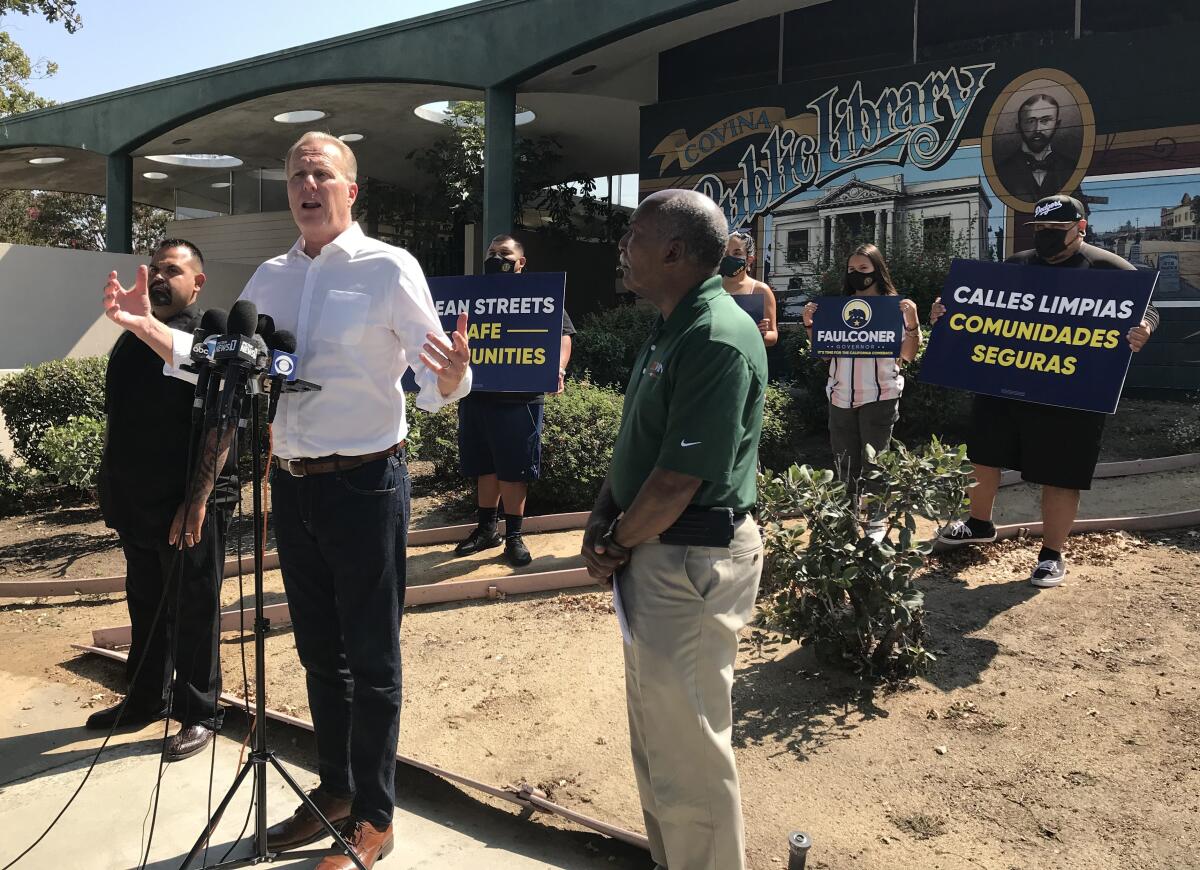 Former San Diego Mayor Kevin Faulconer stands in front of microphones with two people nearby.