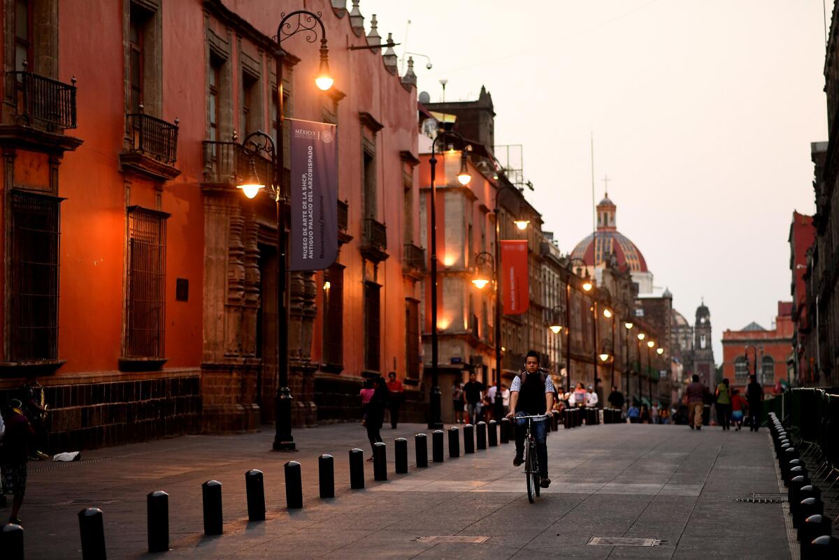 A man rides a bicycle down a street next to a row of buildings in Mexico City.