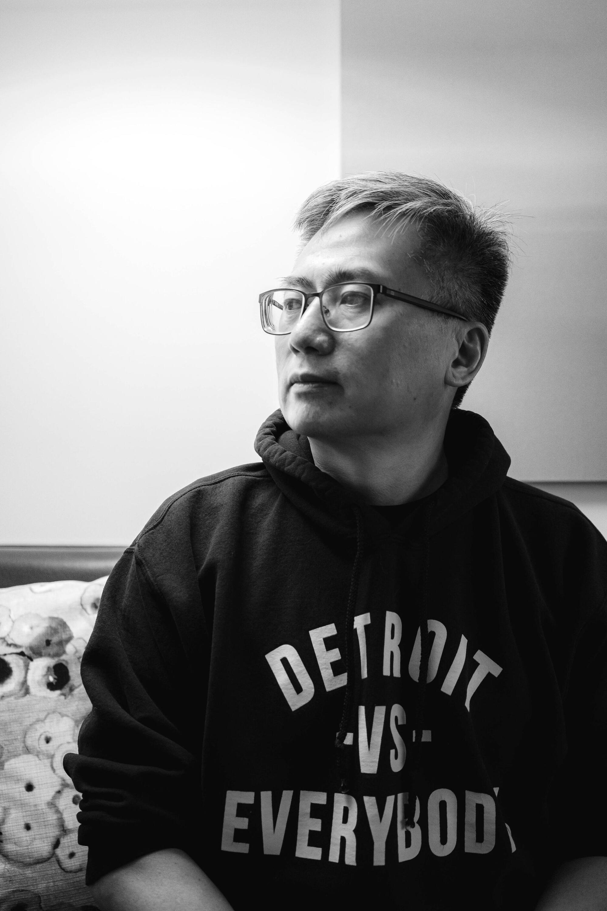 A black and white photo of a man wearing glasses and a hoodie with the words Detroit vs. All