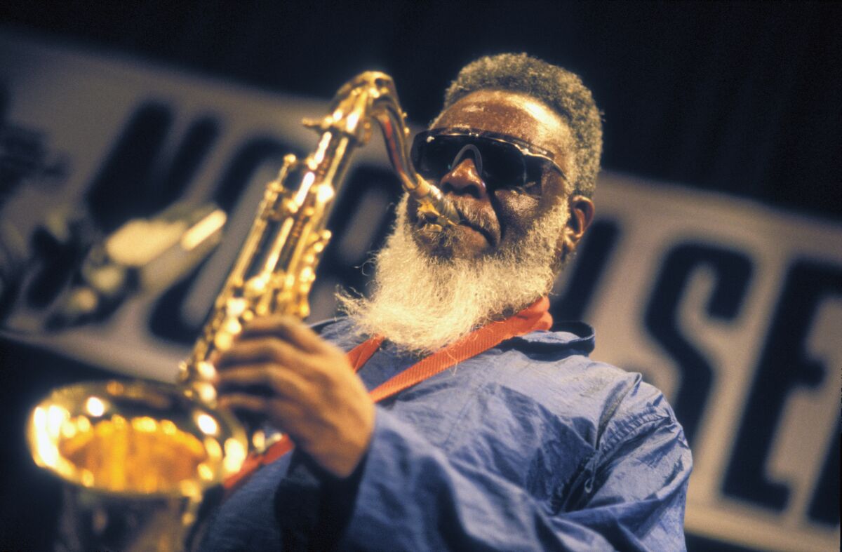 A man with a full white beard and black sunglasses plays the saxophone