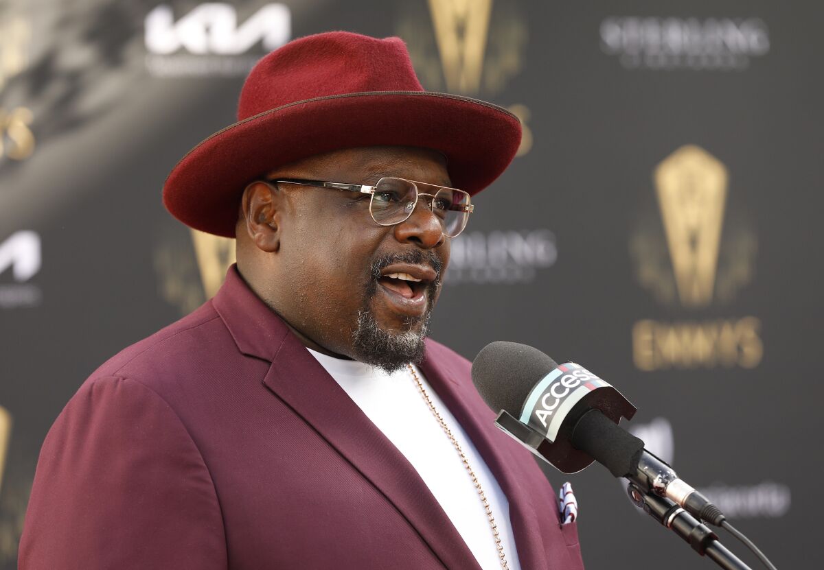 Cedric the Entertainer, wearing a purple suit and hat, speaks into a microphone 