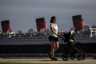 Long Beach, CA - May 25: A woman walks with a stroller along Shoreline Aquatic Park, with the Queen Mary ship in the distance, docked in Long Beach, CA, photographed Tuesday, May 25, 2021. The ship has been a tourist destination and hotel for years and is now in danger of capsizing according to a recent inspection report. (Jay L. Clendenin / Los Angeles Times)