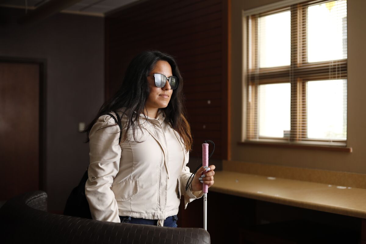 A woman with long hair, wearing sunglasses and holding a mobility cane, stands in front of a window.