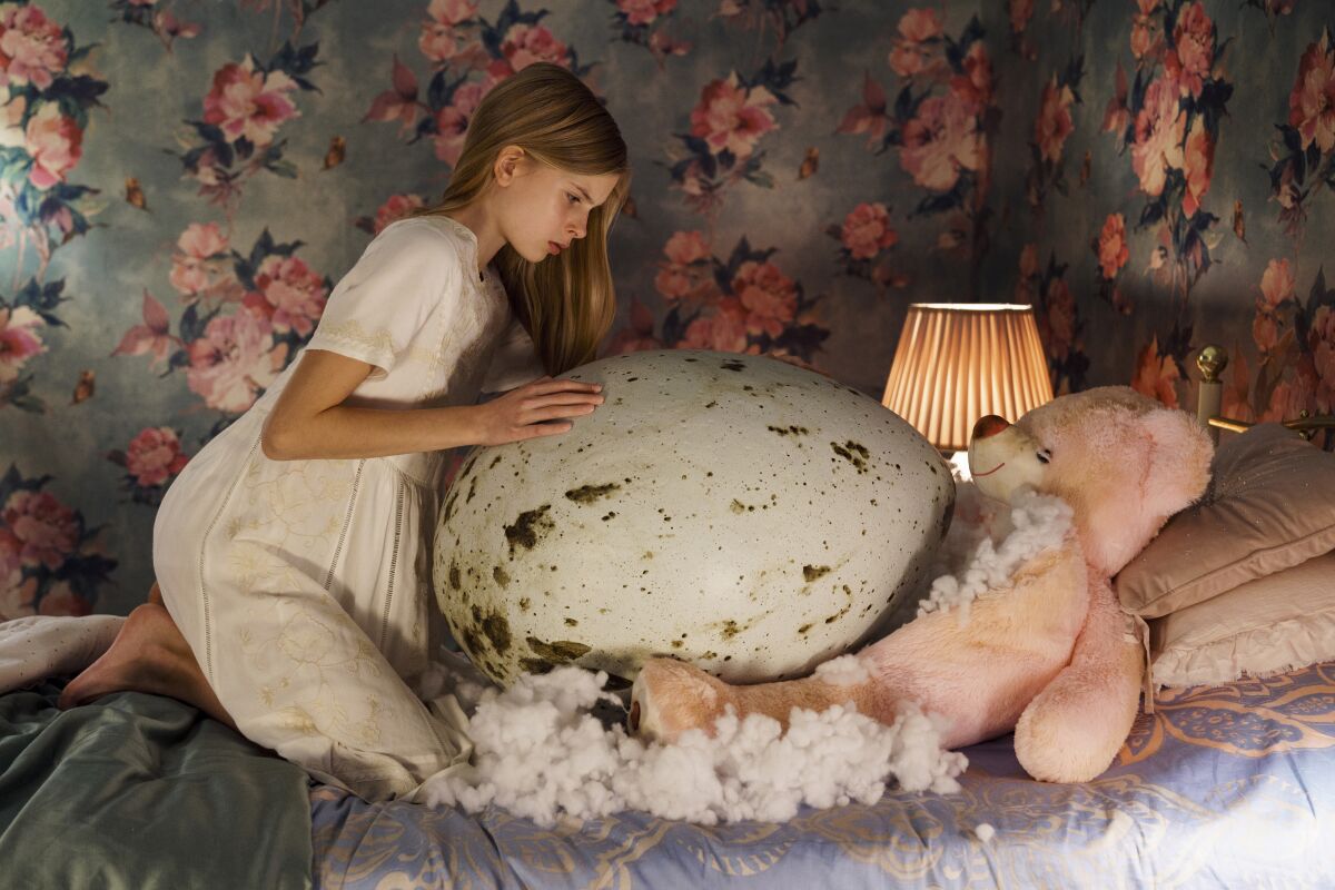 In a bedroom with floral wallpaper, a girl kneels on a bed over a huge egg.