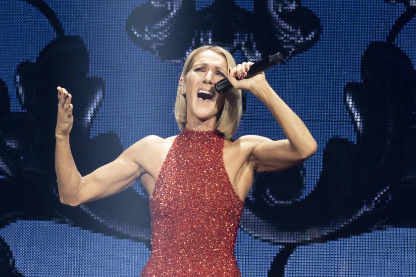 Celine Dion wearing a sparkling red gown sings onstage with her mouth wide open and her arms raised