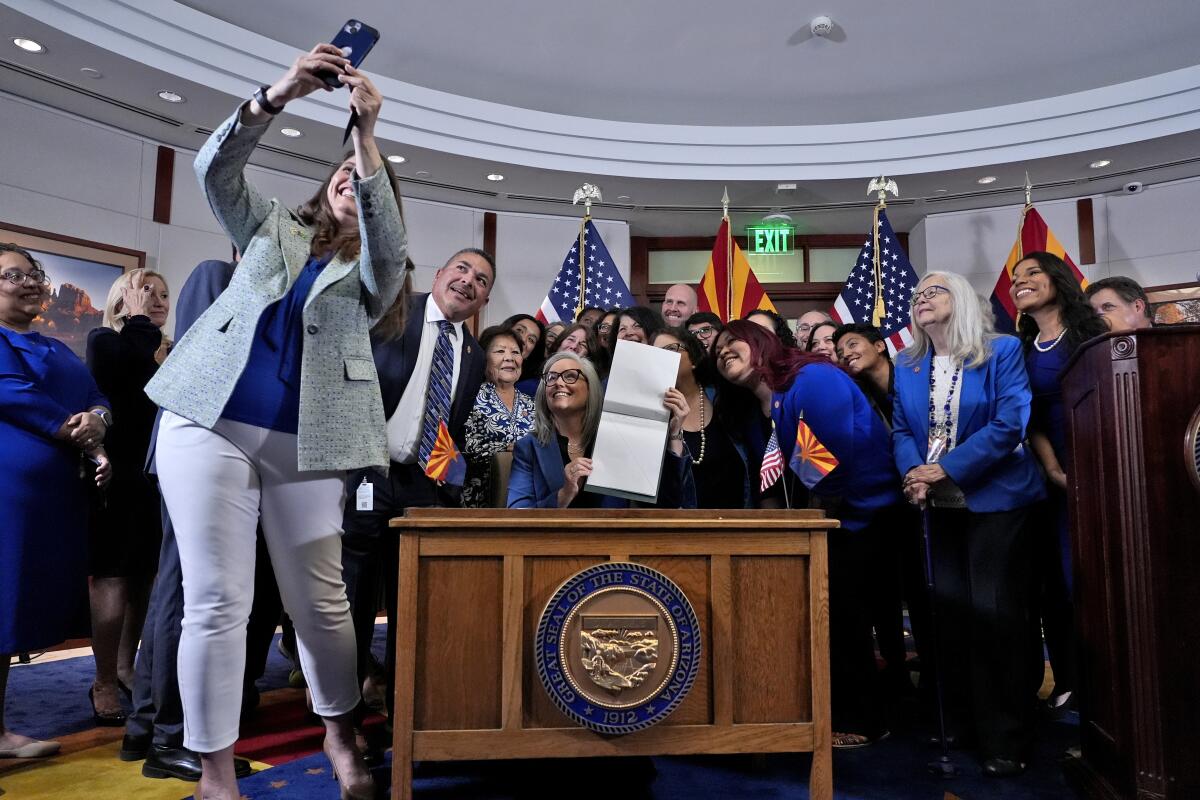 A woman takes a selfie with Arizona Gov. Katie Hobbs and several people standing around her as she sits holding a document.