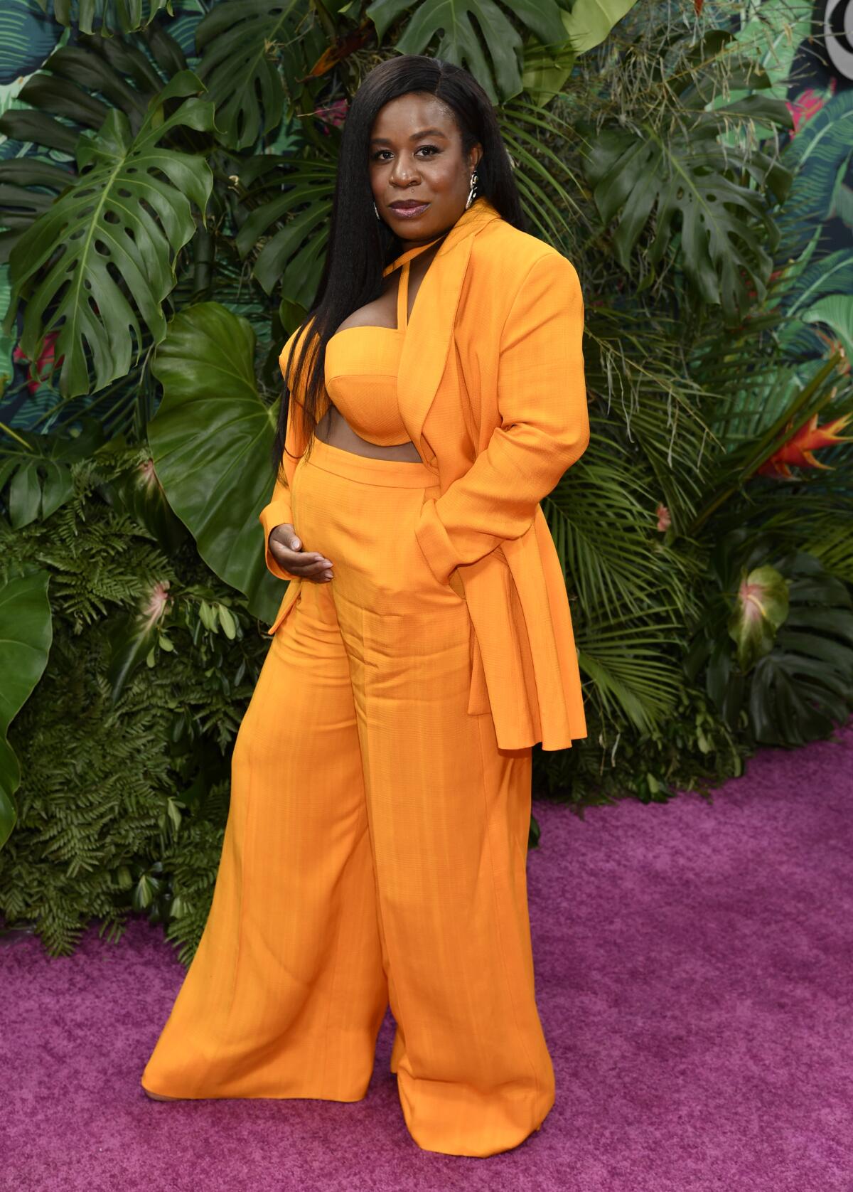 Uzo Aduba poses on a purple carpet cradling her pregnant belly while wearing an orange suit