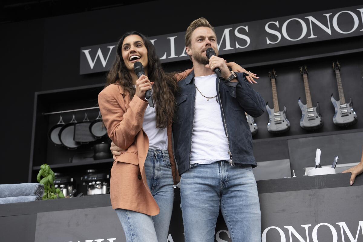 Hayley Erbert and Derek Hough smile and embrace in jeans and casual jackets while holding mics to their mouths