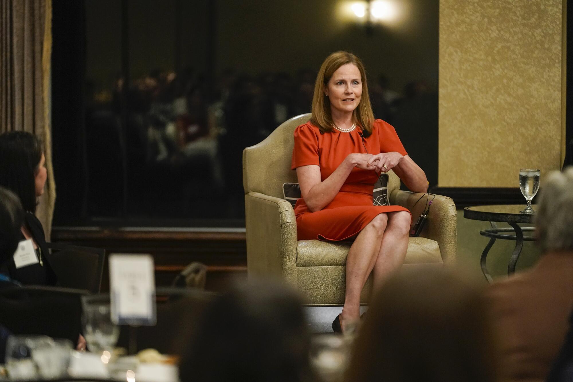 Justice Amy Coney Barrett, in an orange-red dress, sits and addresses a small group, reflected in the window behind her
