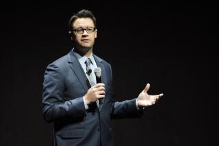 A man with short brown hair wearing a blue suit, speaking into a microphone and gesturing with his hand