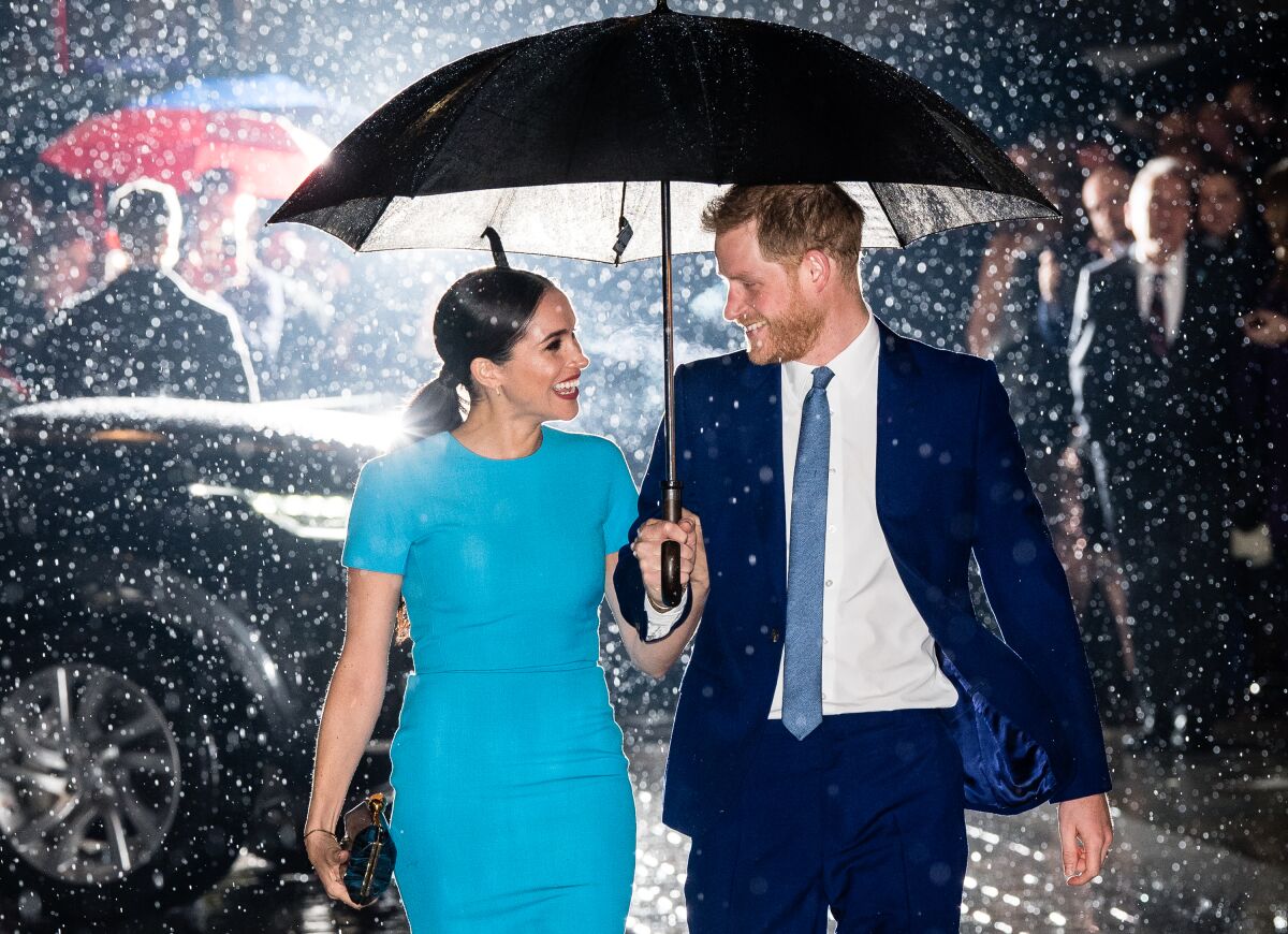 Prince Harry holding an umbrella over himself and Megan on a rainy night, with a car and other pedestrians in the background.