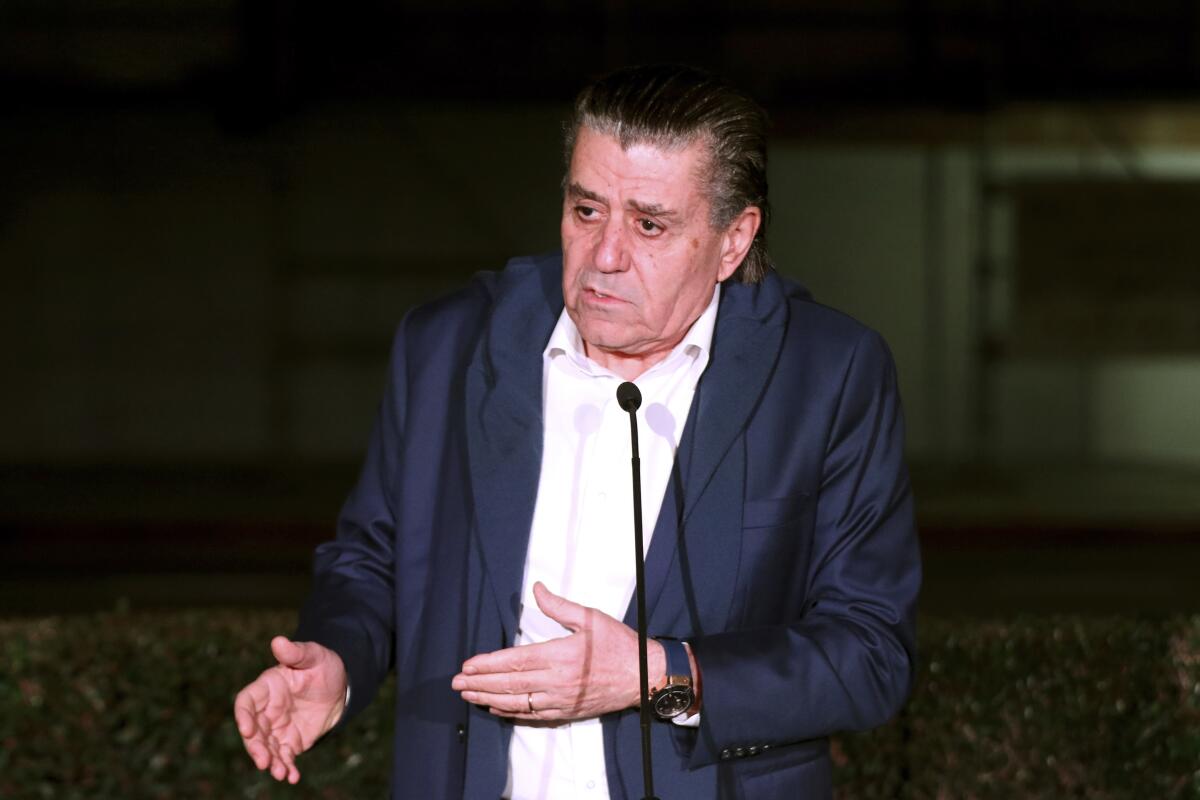 A man in a dark jacket and white shirt speaks at a microphone 