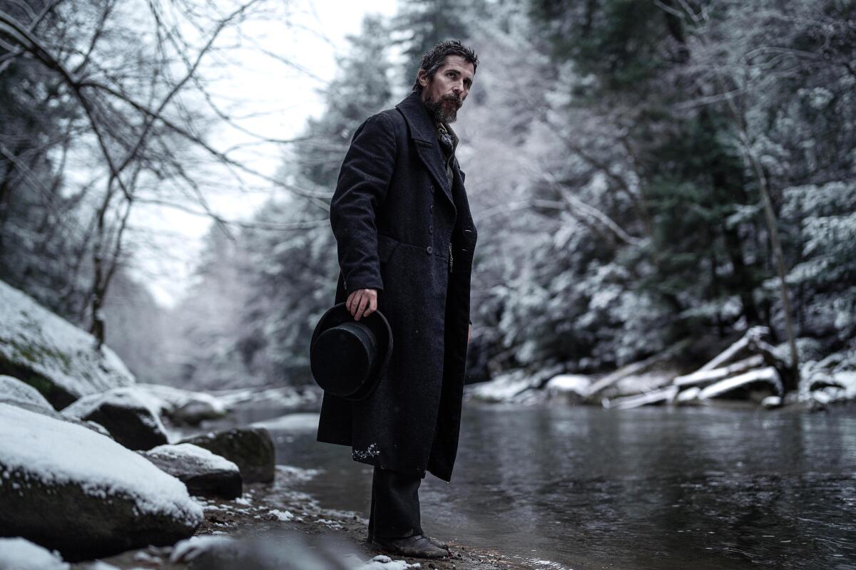 A man in a long coat stands along a snowy river bank.