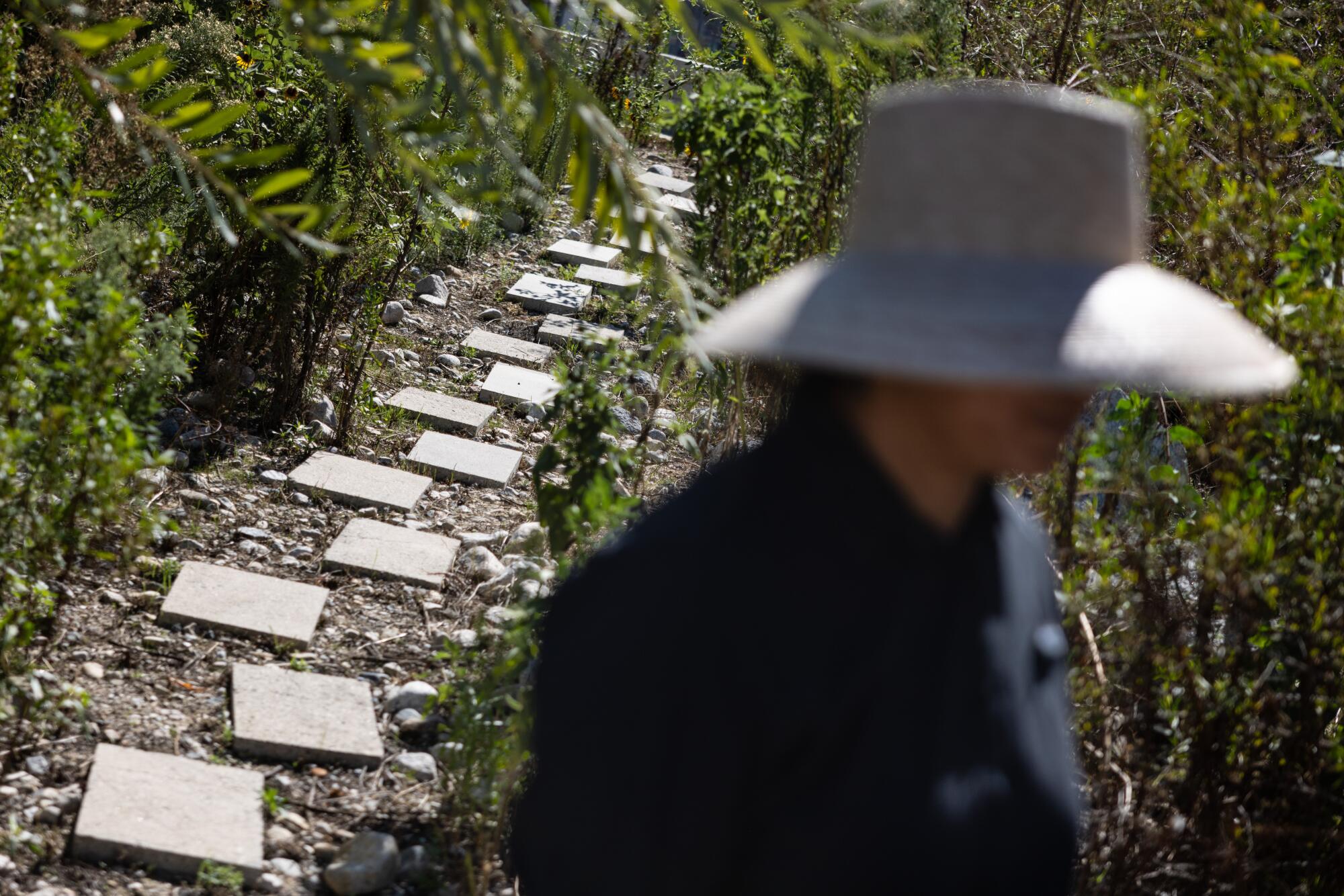 A person in a big gardener's hat walks before a pathway leading into a wooded garden